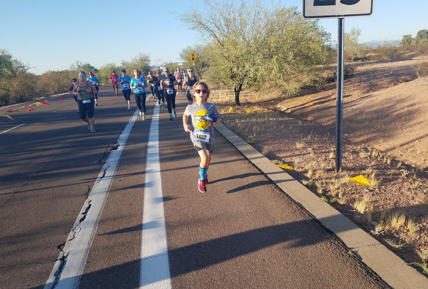 A young runner in sunglasses leading a pack of adults at a 5K race in the desert